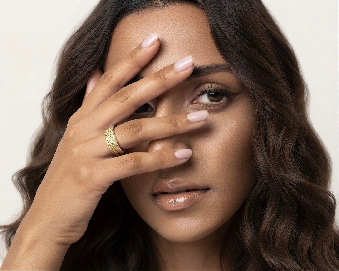 A close-up portrait of a woman with wavy dark brown hair, wearing a textured gold band ring on her finger, with her hand covering part of her face against a light beige background.